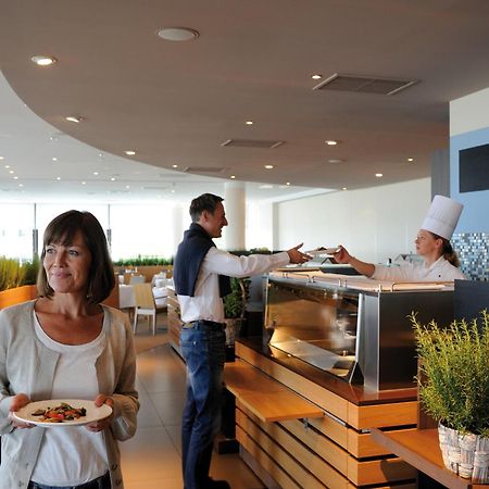 Hotel Neptun Rostock Exterior photo The photo depicts a modern restaurant interior. In the foreground, a woman holding a plate of food looks contemplatively at something in the distance. In the background, a man is interacting with a chef, who is wearing a white chef's hat and is servi