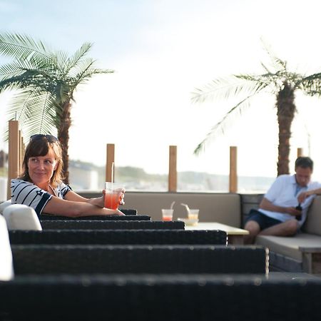 Hotel Neptun Rostock Exterior photo The photo shows a relaxed outdoor setting, likely on a terrace or patio. In the foreground, a woman with dark hair is sitting at a table, holding a colorful drink and looking directly at the camera. She appears to be enjoying her time. In the backgro