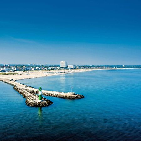 Hotel Neptun Rostock Exterior photo The photo showcases a serene coastal scene. In the foreground, there is a jetty extending into calm, blue waters, marked by a small green lighthouse at its end. The background features a sandy beach with a clear sky overhead, indicating a sunny day. 