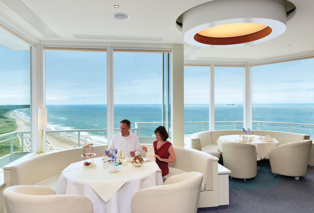 Hotel Neptun Rostock Exterior photo The photo shows a restaurant setting with large windows that offer a view of the sea. Inside, there is a dining area with modern, light-colored furniture, including round tables and comfortable seating. Two people are sitting at one of the tables, en