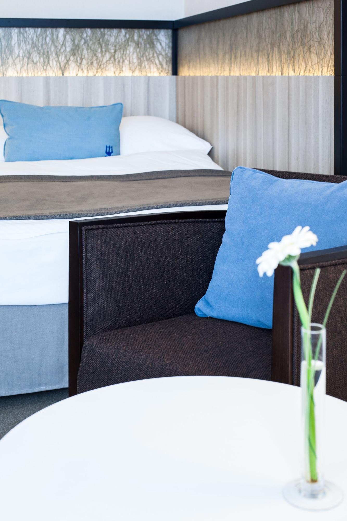 Hotel Neptun Rostock Exterior photo The photo features a modern hotel room setting. In the foreground, there is a dark-colored chair with a light blue cushion. A small white table is in front of the chair, holding a decorative vase with a single flower. In the background, a neatly made