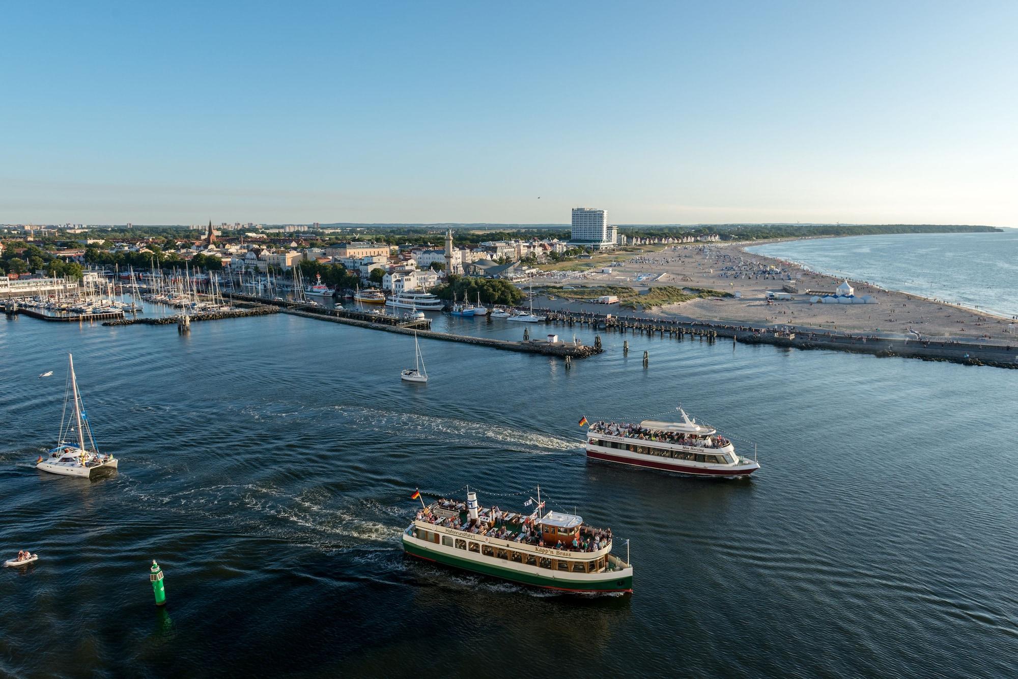 Hotel Neptun Rostock Exterior photo The photo depicts a coastal scene featuring a harbor or marina. There are several boats navigating through the water, with at least two larger vessels evident, one possibly a ferry, both creating wake trails behind them. In the background, there are 