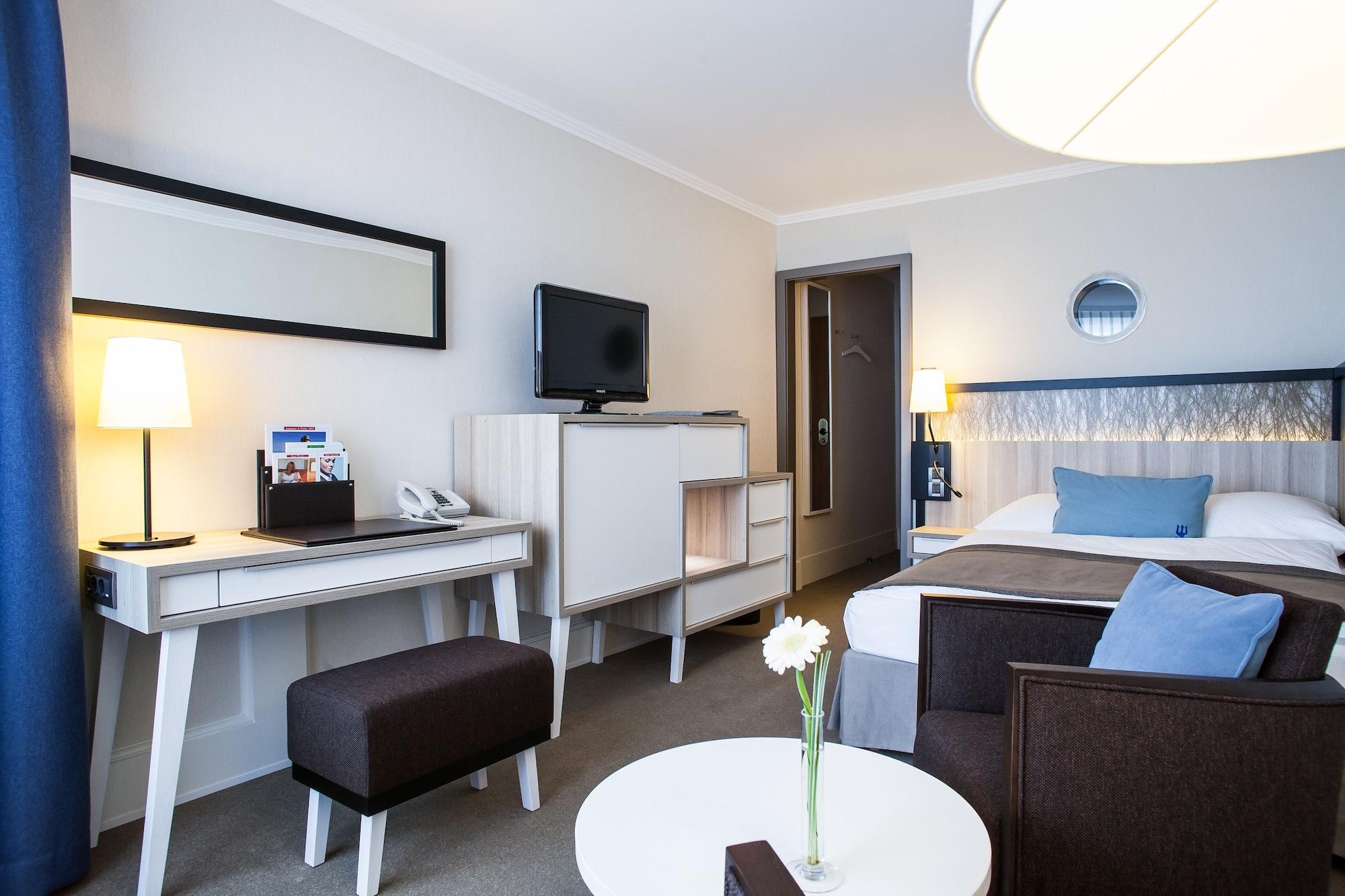 Hotel Neptun Rostock Exterior photo The photo shows a modern hotel room. It features a neatly made bed with a couple of blue pillows, a desk area with a lamp, and a flat-screen television mounted on the wall. There's a small round table with a flower in the center and a dark-colored ar