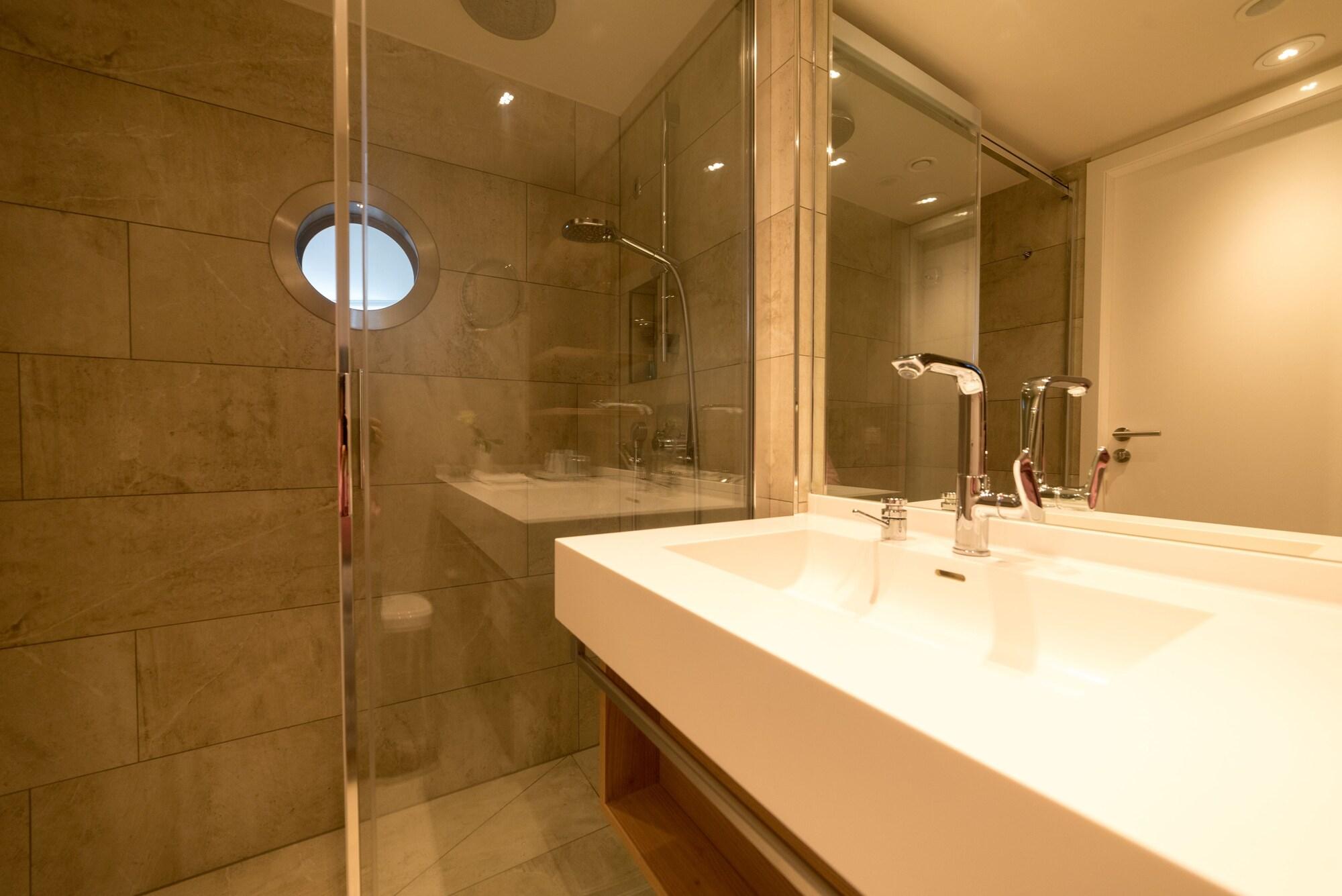 Hotel Neptun Rostock Exterior photo The photo shows a modern bathroom featuring a sleek design. There is a glass shower enclosure on one side, with a showerhead visible. Adjacent to the shower is a large, white countertop sink with two faucets. The walls are tiled with light-colored st