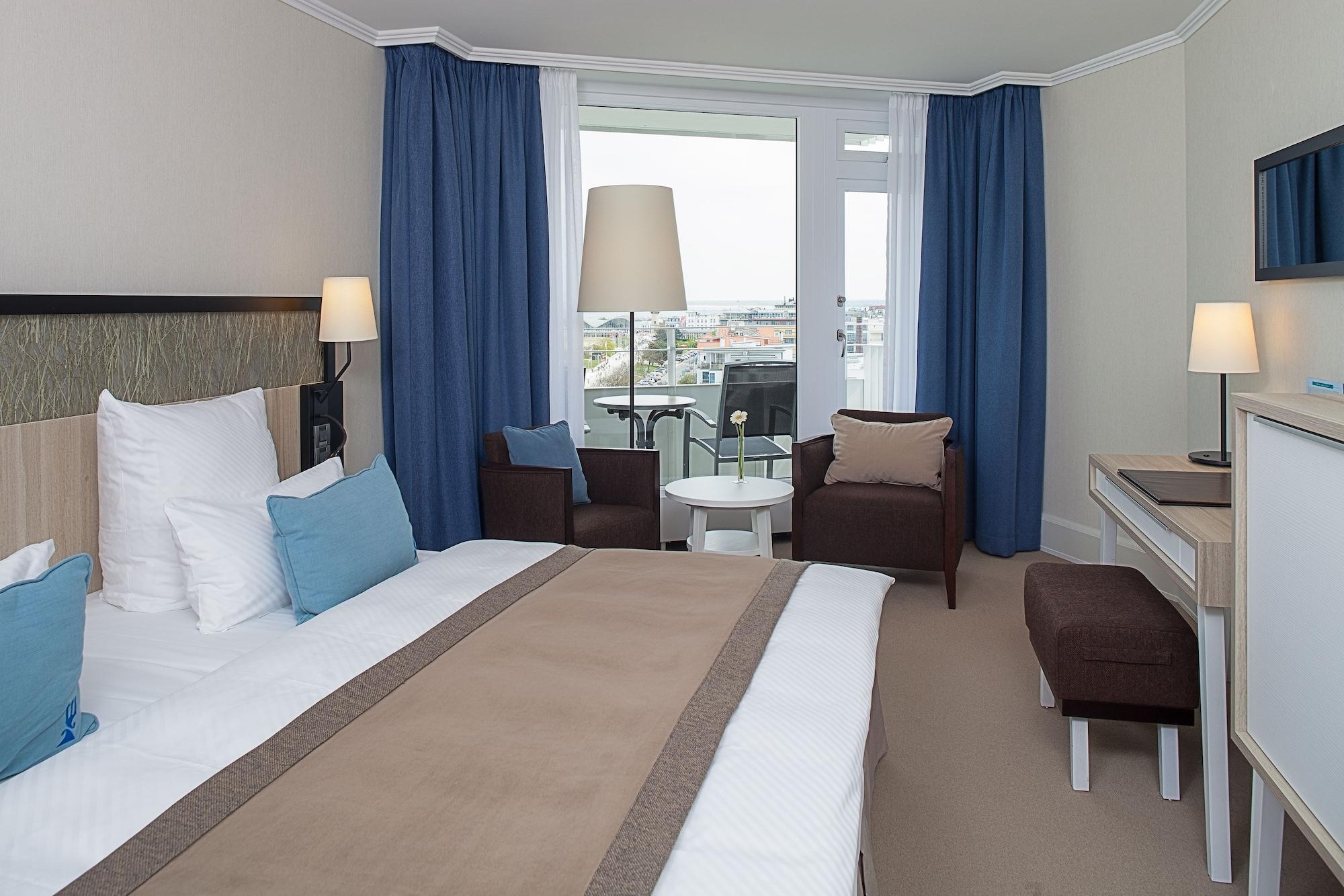 Hotel Neptun Rostock Exterior photo The photo shows a modern hotel room featuring a neatly made bed with white sheets and a beige throw blanket. There are two blue pillows on the bed. The room has large windows with light blue curtains that allow natural light to enter, and a lamp is p