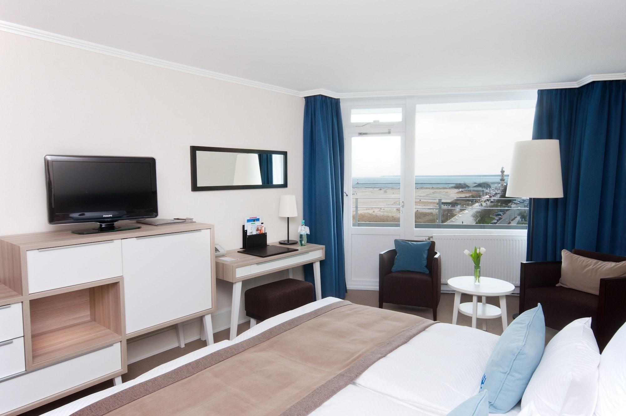 Hotel Neptun Rostock Exterior photo The photo shows a modern hotel room. It features a neatly made bed with light-colored linens. Opposite the bed is a wall-mounted flat-screen TV, accompanied by a console and a mirror. There is a small desk area with a chair, and next to it, a round t