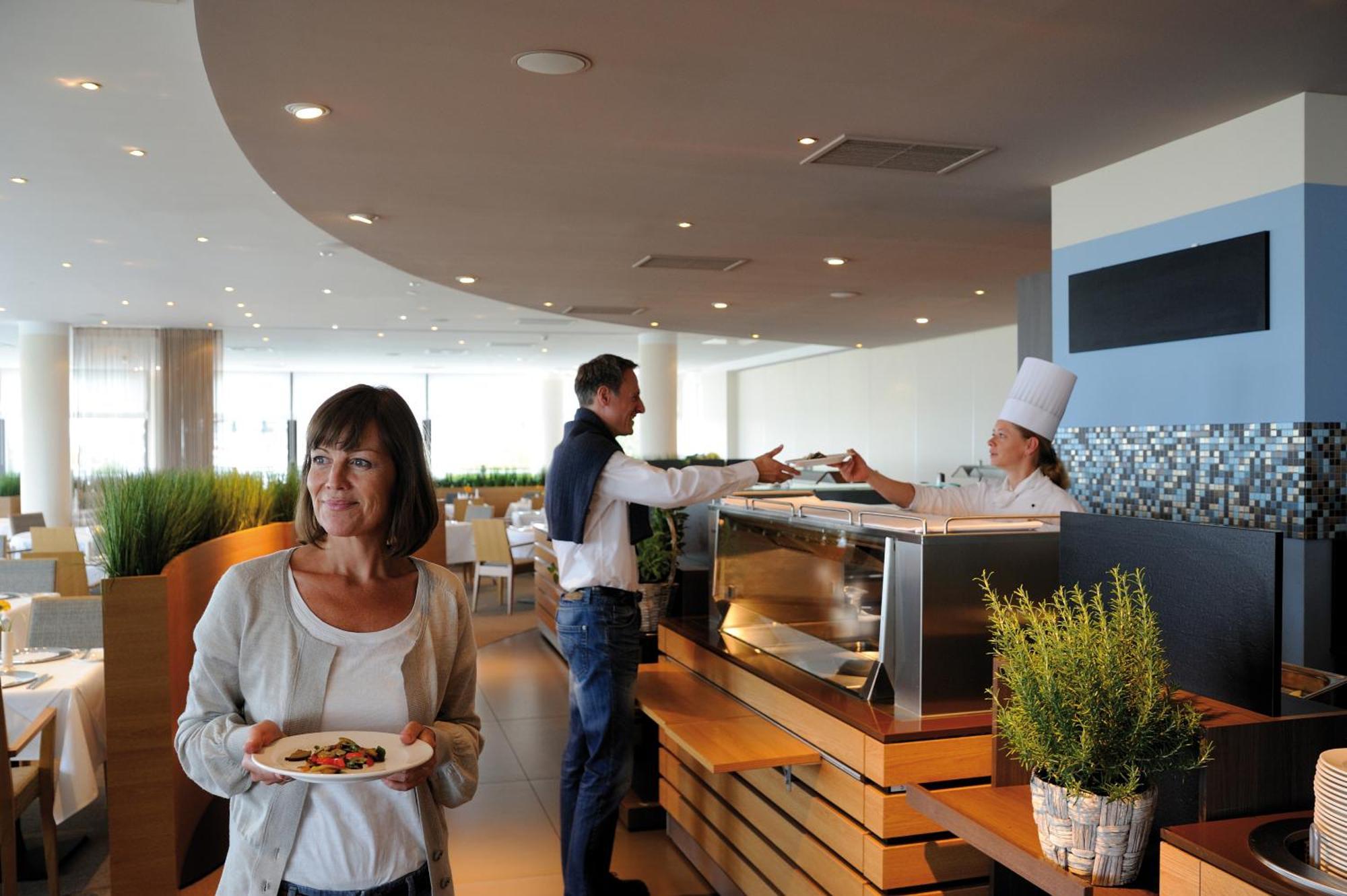 Hotel Neptun Rostock Exterior photo The photo depicts a modern restaurant interior. In the foreground, a woman holding a plate of food looks contemplatively at something in the distance. In the background, a man is interacting with a chef, who is wearing a white chef's hat and is servi