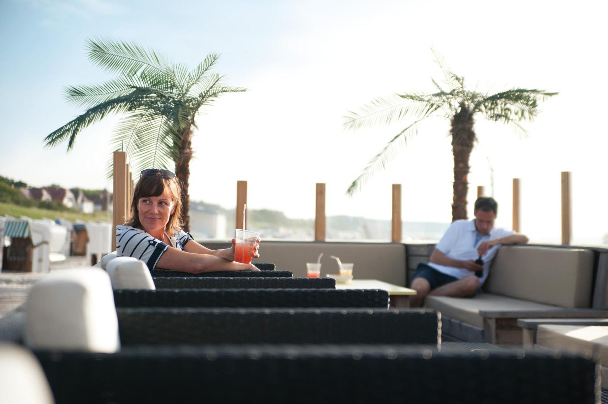 Hotel Neptun Rostock Exterior photo The photo shows a relaxed outdoor setting, likely on a terrace or patio. In the foreground, a woman with dark hair is sitting at a table, holding a colorful drink and looking directly at the camera. She appears to be enjoying her time. In the backgro