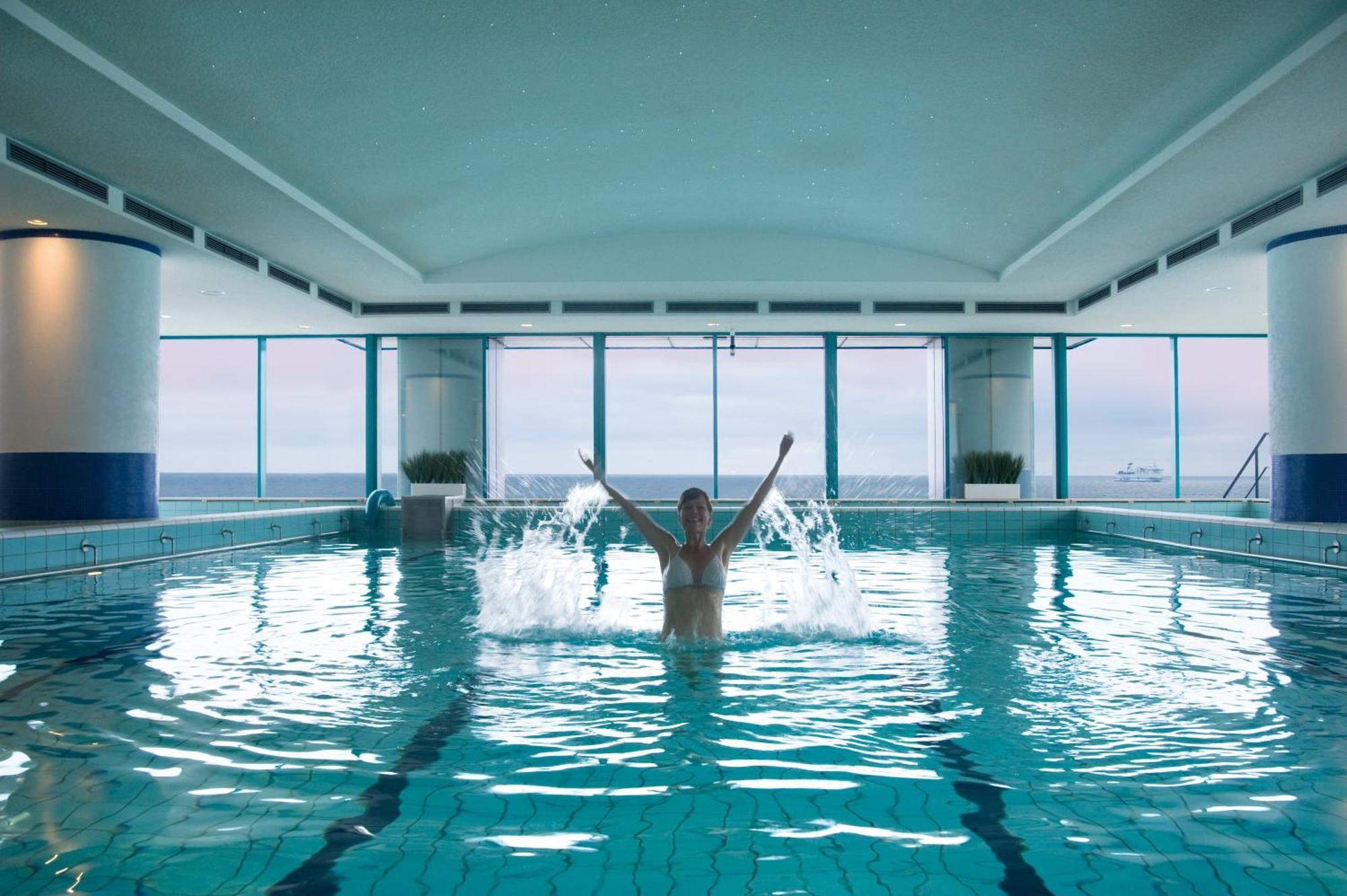 Hotel Neptun Rostock Exterior photo The photo shows an indoor swimming pool with large windows that offer a view of the ocean. In the center of the pool, a person is standing in the water with their arms raised, creating splashes. The serene environment is enhanced by the soft lighting