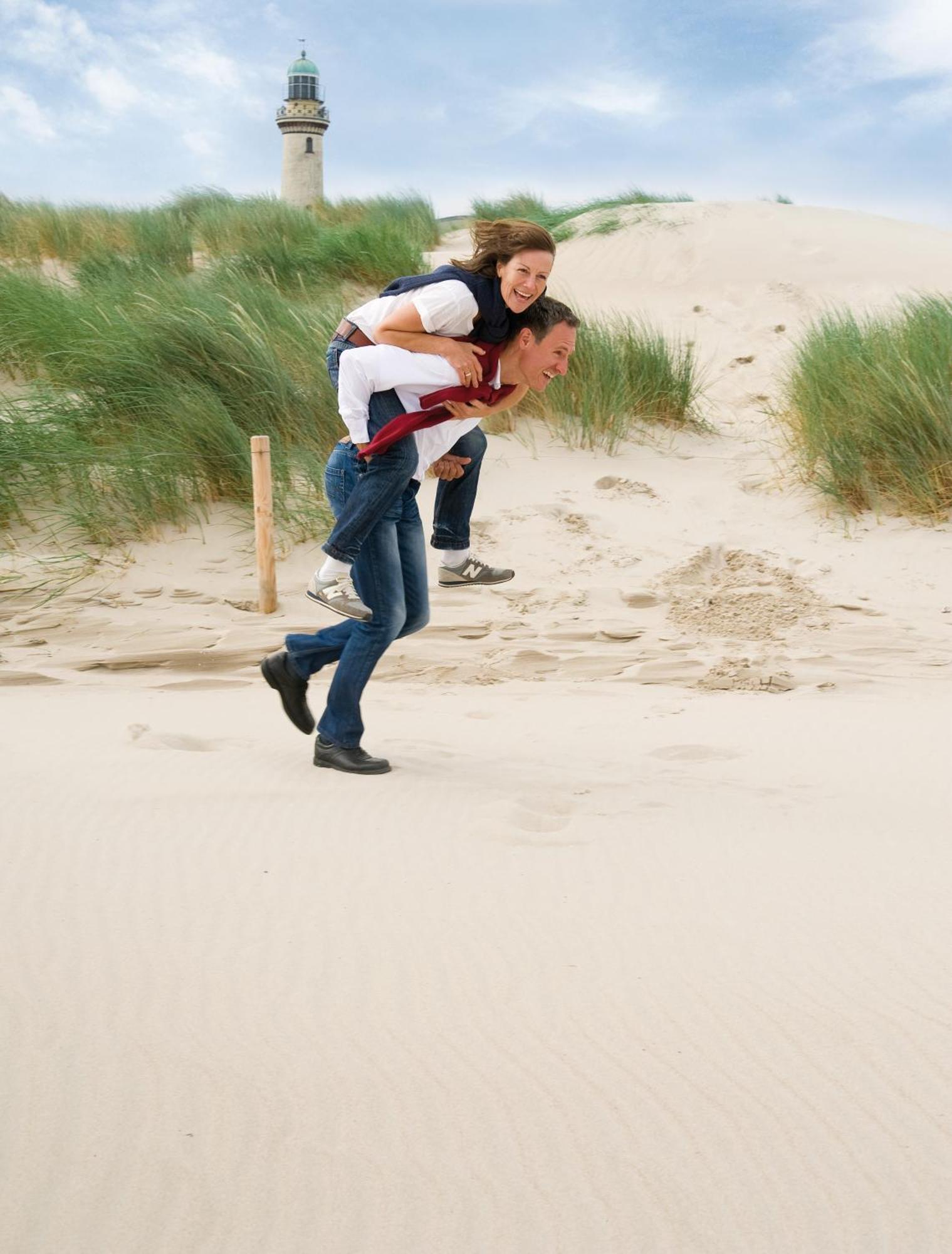 Hotel Neptun Rostock Exterior photo The photo shows two people enjoying a playful moment on a sandy area, likely a beach or dune. One person is carrying the other on their back, running with a joyful expression. The person being carried appears to be having fun, possibly laughing or sm