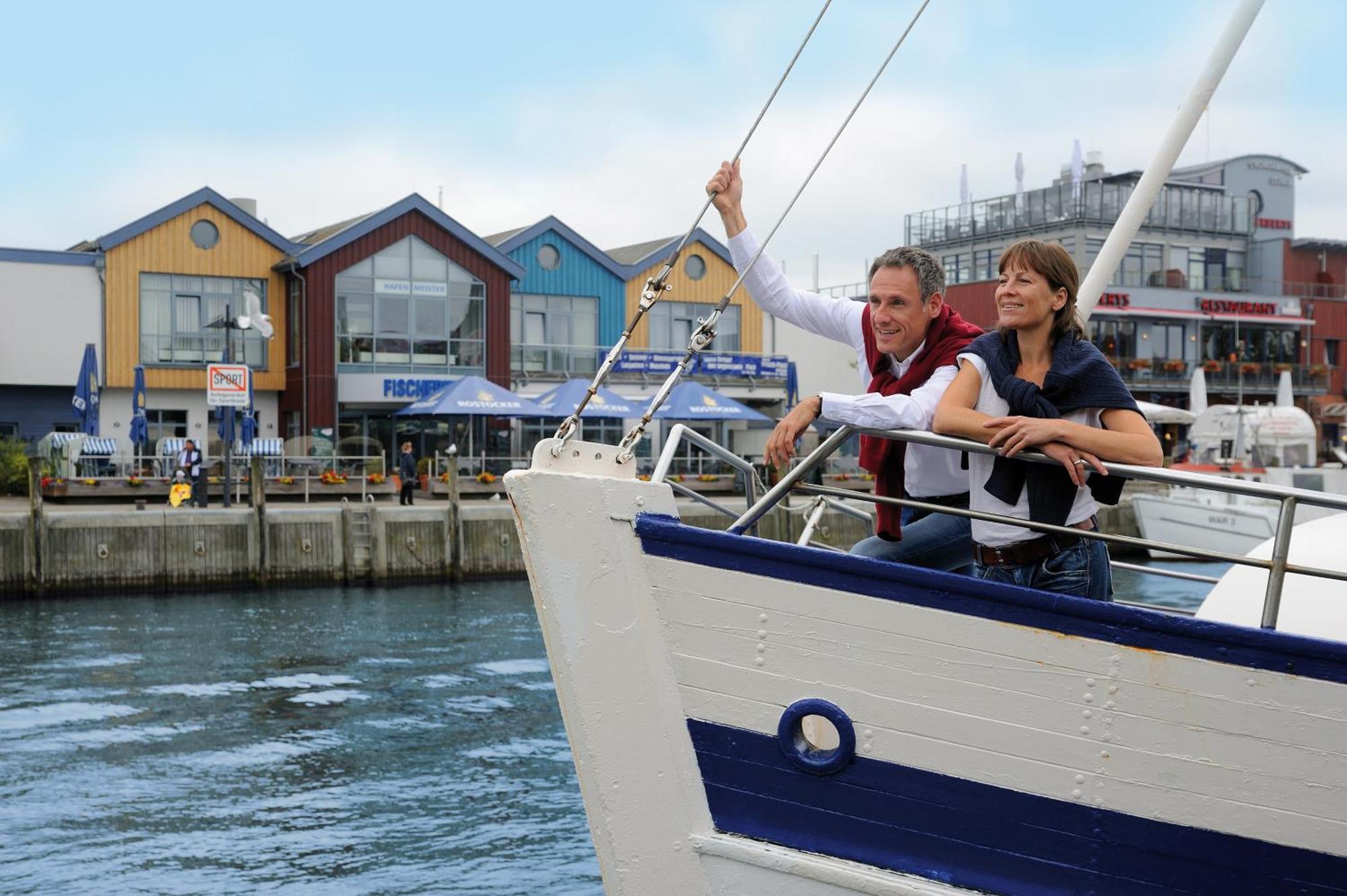 Hotel Neptun Rostock Exterior photo The photo shows a couple standing on the bow of a boat in a harbor. The man is holding onto a railing, looking out at the water, while the woman stands close to him, enjoying the view. In the background, there are colorful buildings by the waterfront