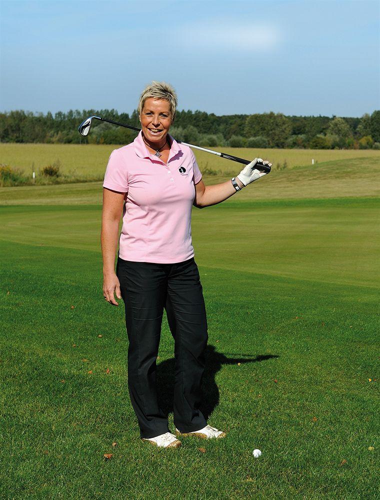 Hotel Neptun Rostock Exterior photo The image shows a woman standing on a golf course, dressed in a pink polo shirt and black pants. She is holding a golf club over her shoulder with one hand and smiling at the camera. The background features a grassy area and trees, indicating a sunny