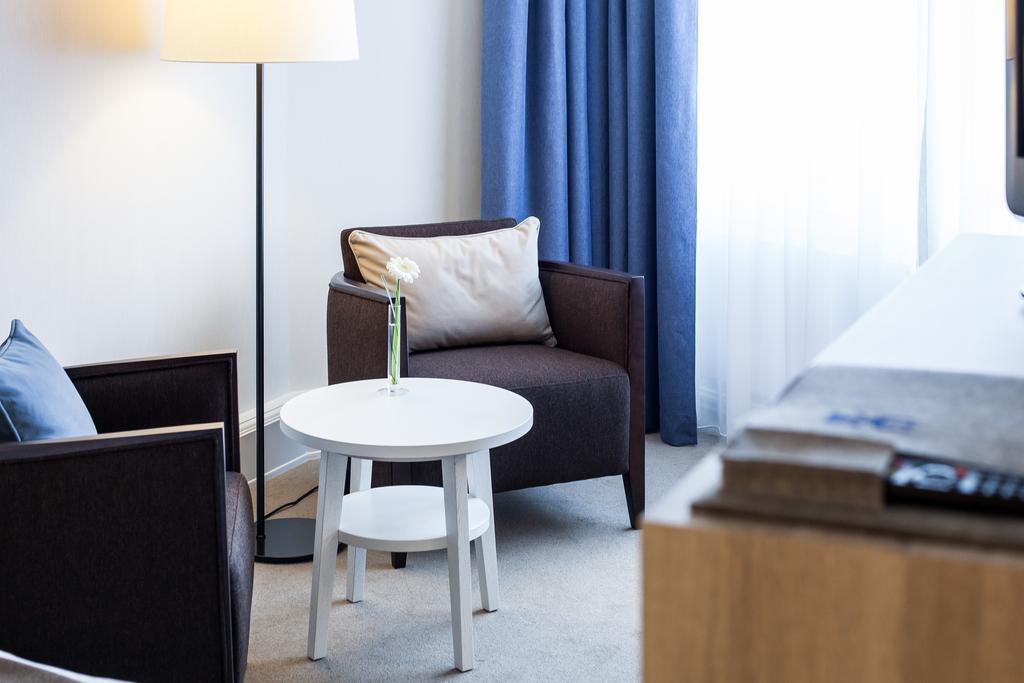 Hotel Neptun Rostock Exterior photo The photo shows a cozy corner of a room featuring two armchairs—one dark brown and the other light blue. Between them is a small white table with a flower in a vase on top. There's a floor lamp next to the brown chair, and a set of neutral-colored cu