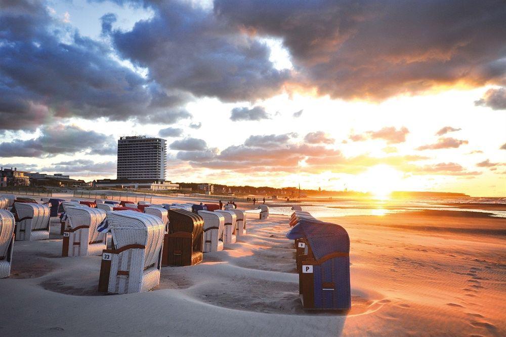 Hotel Neptun Rostock Exterior photo The photo shows a beach scene during sunset. In the foreground, there are several colorful beach huts or chairs arranged in a row on the sandy beach. The sky is filled with dramatic clouds, illuminated by the warm colors of the setting sun. In the ba