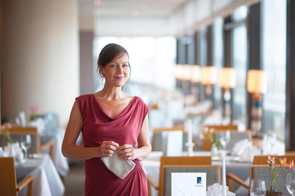 Hotel Neptun Rostock Exterior photo The photo shows a woman standing in a stylish restaurant. She is wearing a red dress and holding a small clutch. The background features a well-lit dining area with tables set for meals, including white tablecloths, elegant dinnerware, and decorative