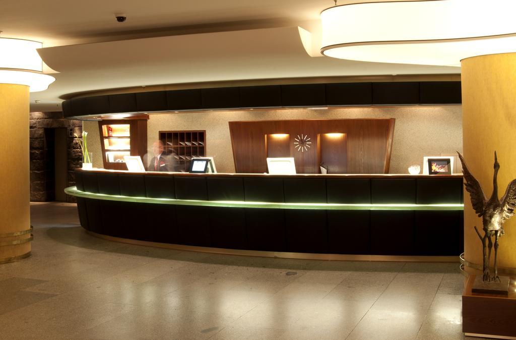 Hotel Neptun Rostock Exterior photo The image shows a modern hotel reception area. The reception desk is curved and features a sleek design with a dark wood finish and a lighted strip along the bottom. Behind the desk, there are shelves and a decorative wall with a clock on it. The lig