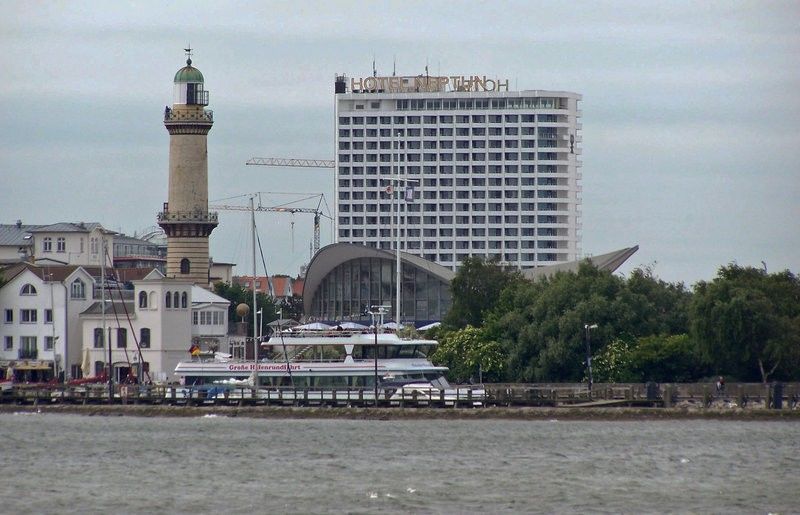 Hotel Neptun Rostock Exterior photo The photo shows a waterfront scene featuring a combination of buildings and a yacht. On the left, there is a tall, cylindrical lighthouse made of stone, which has a balcony at the top. Next to the lighthouse, there is a more traditional building with