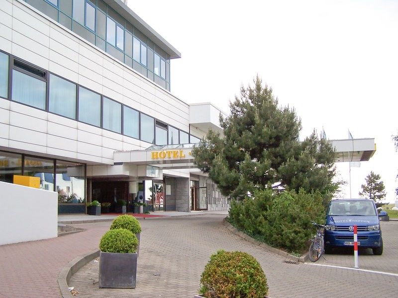 Hotel Neptun Rostock Exterior photo The photo shows the exterior of a hotel building. The structure features a modern design with large windows and a sign prominently displaying the word "HOTEL." In front of the hotel, there are landscaped areas with plants and shrubs. A blue car is pa