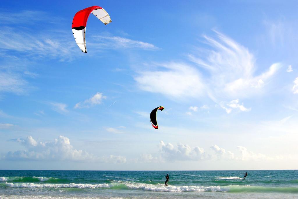 Hotel Neptun Rostock Exterior photo The photo shows a beach scene with two kite surfers riding the waves. They are being pulled by colorful kites—a red and black one, and a yellow and black one—soaring high in the blue sky. The ocean appears calm with gentle waves, and the sky is partl