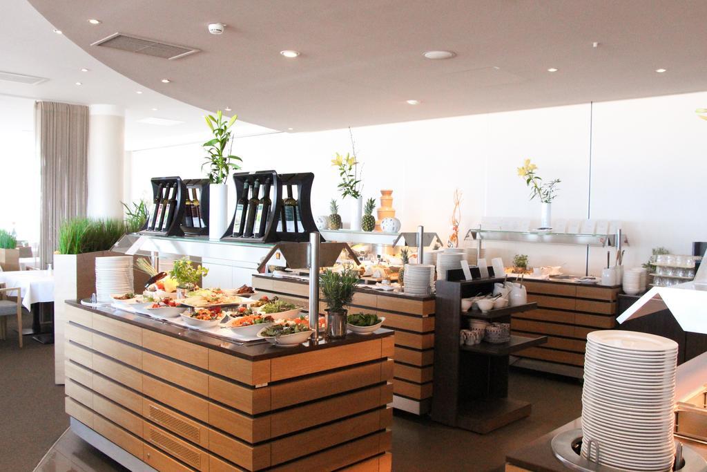 Hotel Neptun Rostock Exterior photo The photo depicts a modern buffet setup in a bright, well-lit dining area. There are several wooden serving stations arranged neatly, each featuring a variety of food items. The buffet includes salads and other dishes, presented in an organized manne