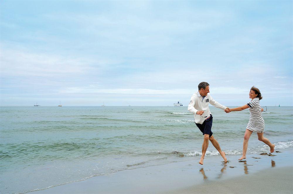 Hotel Neptun Rostock Exterior photo The photo shows a couple running along the beach, holding hands. The scene is set by the ocean, with gentle waves lapping at the shore. The sky is partly cloudy, creating a calm and serene atmosphere. The man is wearing a light-colored long-sleeve sh