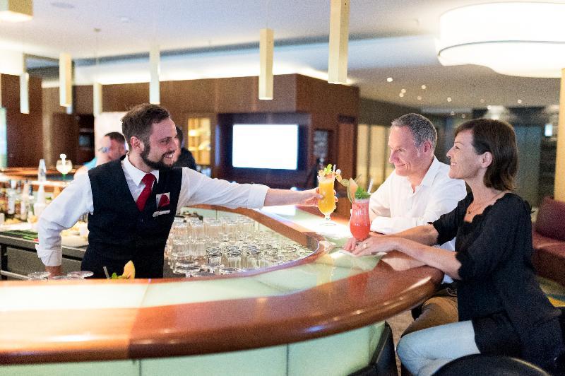 Hotel Neptun Rostock Exterior photo The photo shows a bartender serving colorful cocktails to two customers at a bar. The bartender, dressed in a black vest and white shirt, is handing over drinks to a man and a woman who are seated at the bar. The setting appears to be a modern and up