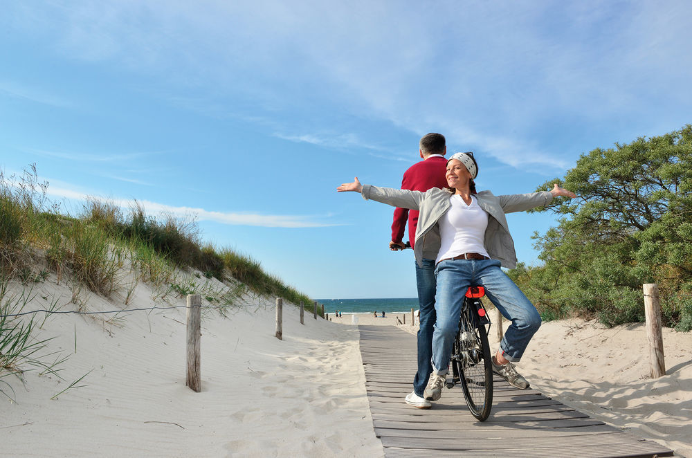 Hotel Neptun Rostock Exterior photo The photo depicts a scenic beach path with two people enjoying their time outdoors. One person is riding a bicycle on a wooden walkway, while the other is seated on the bike, facing backward with arms outstretched, conveying a sense of joy and freedo