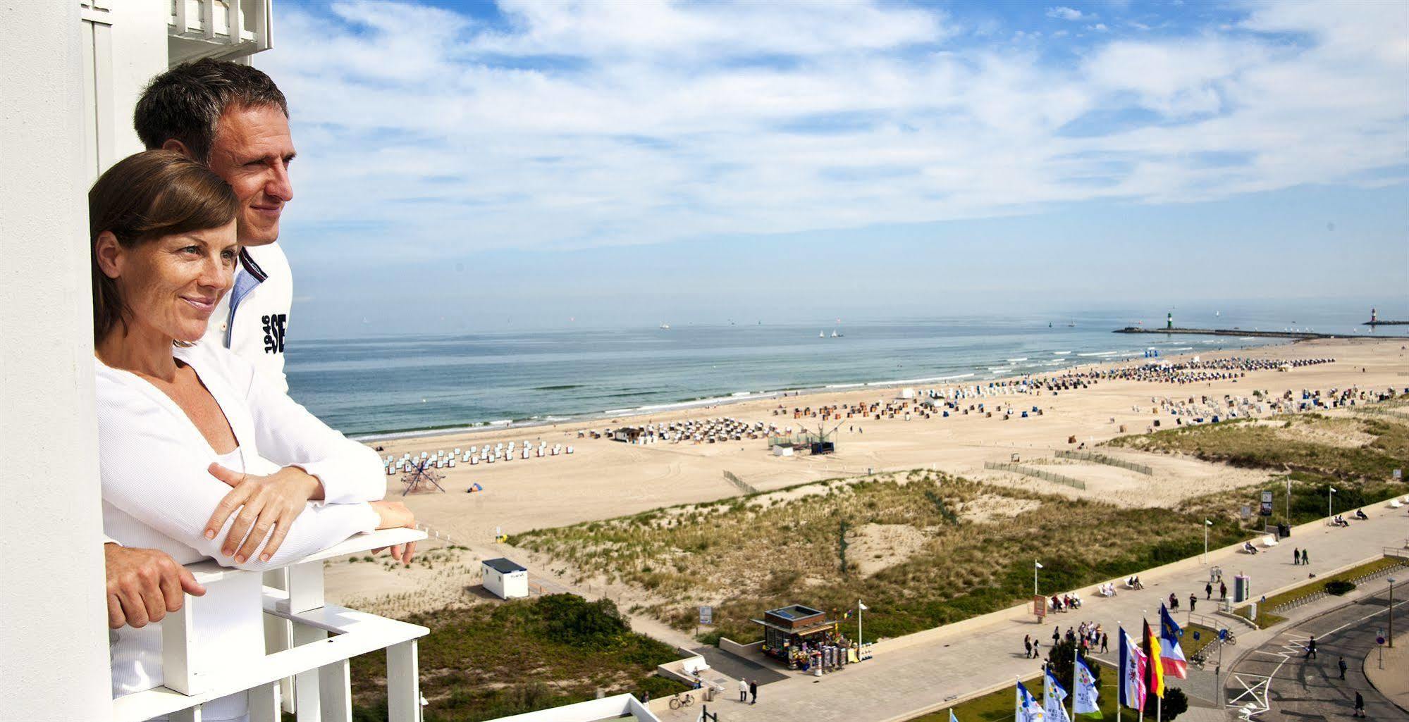 Hotel Neptun Rostock Exterior photo The photo shows a scenic view of a beach with a wide stretch of sand and the ocean in the background. There are numerous beach umbrellas arranged in rows on the sand, suggesting that people are enjoying a day at the beach. In the foreground, a person