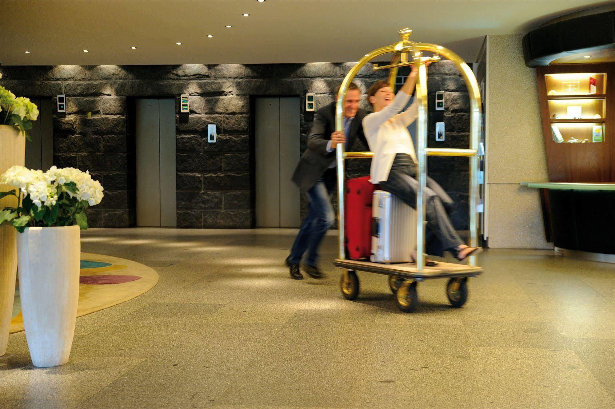 Hotel Neptun Rostock Exterior photo The photo depicts a lively scene in a hotel lobby. A man is playfully pushing a luggage cart, on which a woman is riding. The cart is adorned with a golden frame and contains bags, including a red suitcase and a larger white item. The background feat