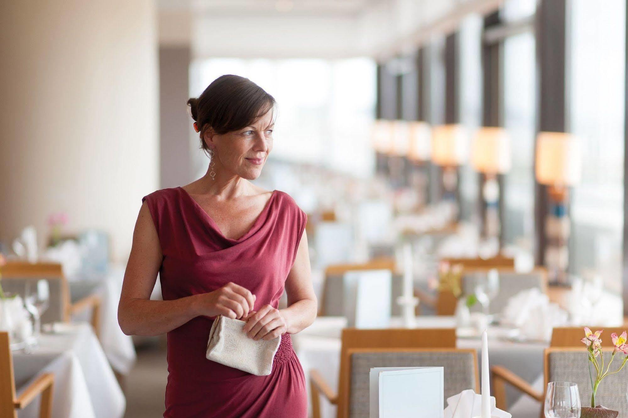 Hotel Neptun Rostock Exterior photo The photo depicts a woman standing in a restaurant setting. She is dressed in a stylish red dress and holding a small purse. The restaurant has an elegant atmosphere, with tables set for dining, featuring white tablecloths and neatly arranged tablewa