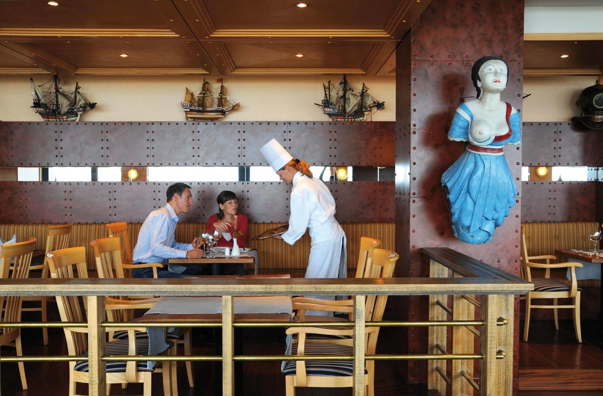 Hotel Neptun Rostock Exterior photo The photo depicts a restaurant setting featuring a couple at a table with a waiter or waitress. The couple appears to be engaged in conversation while being served. The staff member is dressed in a chef’s uniform, complete with a white hat. There are