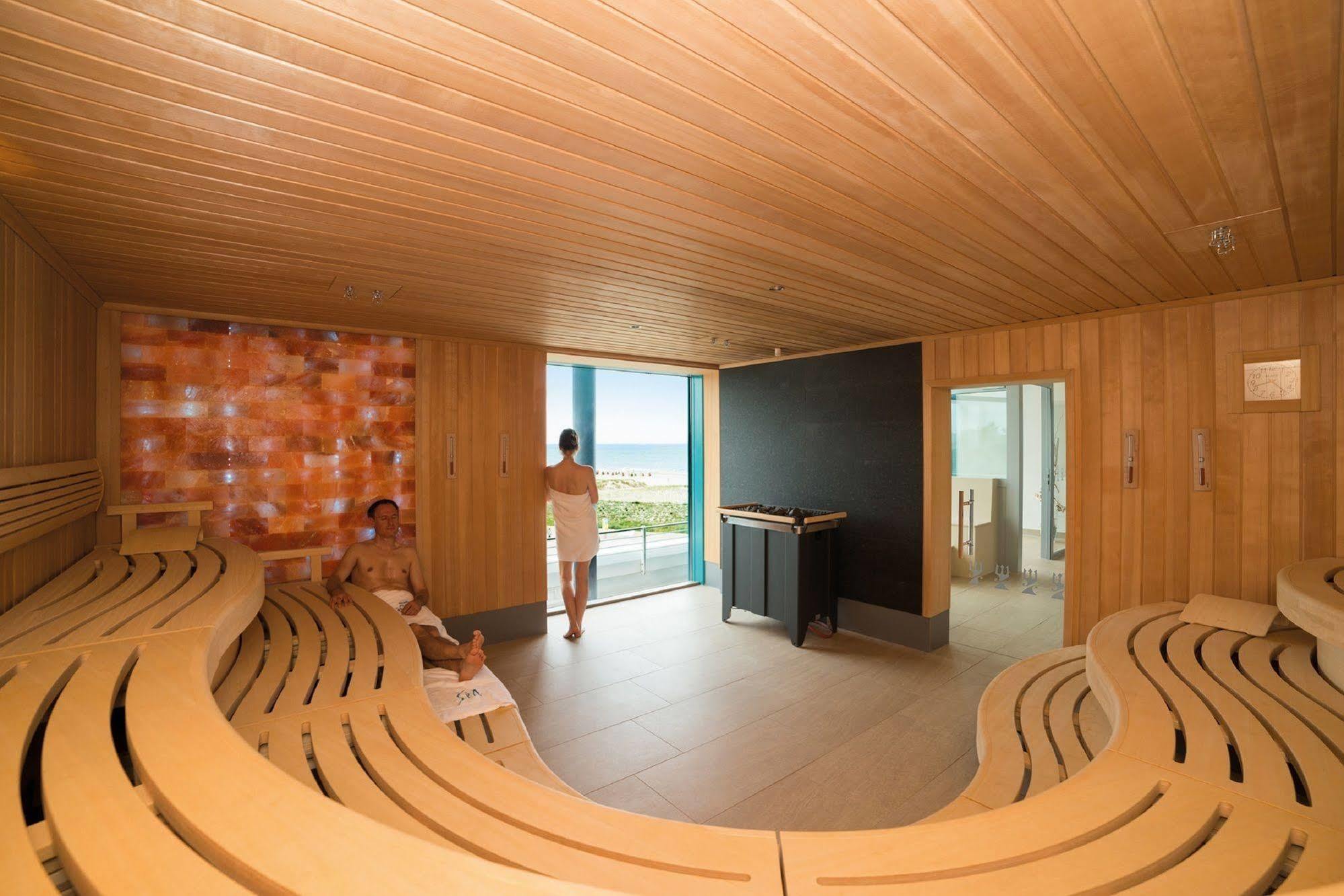 Hotel Neptun Rostock Exterior photo The photo shows an interior of a sauna. The room features a wooden design, with benches arranged in a semicircular layout. There's a large window that offers a view of the outside, allowing natural light to enter the space. One person is standing nea