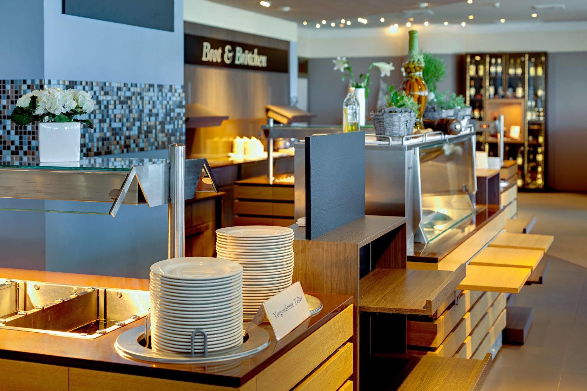 Hotel Neptun Rostock Exterior photo The photo shows a modern dining area, likely a buffet restaurant or a dining hall. There are neatly arranged stacks of white plates on a wooden counter, and various food displays behind glass. The decor appears sleek and contemporary, with well-lit a