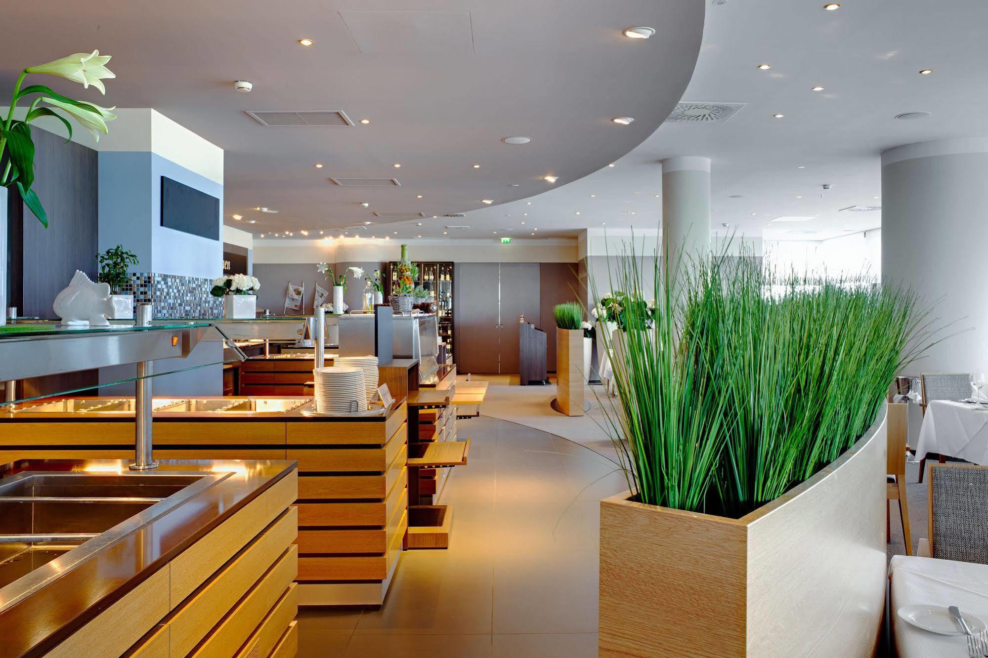 Hotel Neptun Rostock Exterior photo The photo shows a modern restaurant interior featuring a sleek, contemporary design. The space includes wooden elements, likely part of the furnishings, with clean lines and a stylish layout. There are several dining areas, with wooden tables and ben