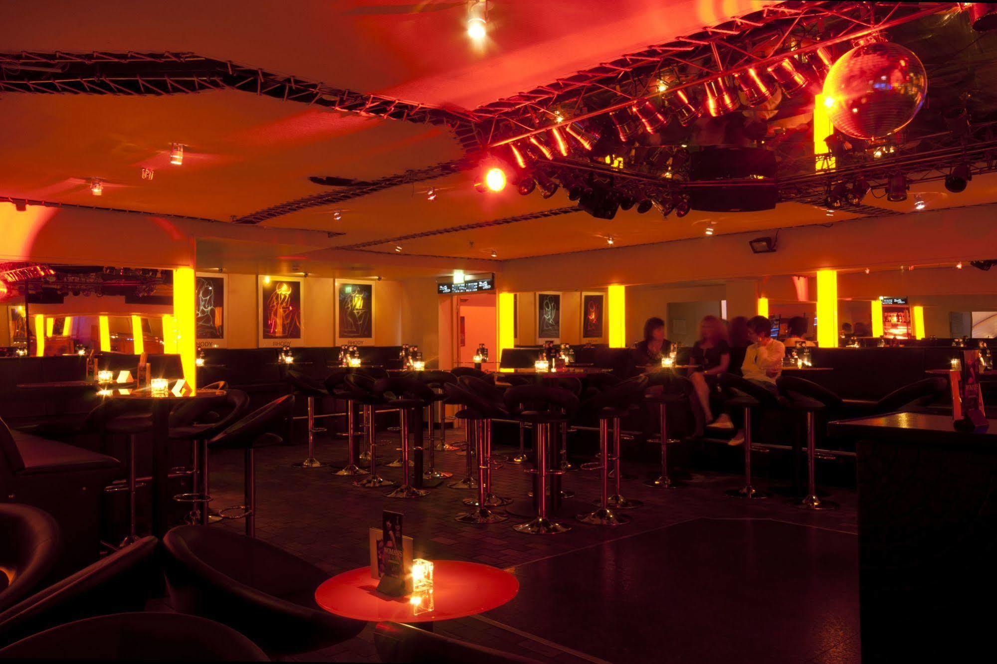 Hotel Neptun Rostock Exterior photo The photo shows the interior of a bar or lounge setting. The space is dimly lit with warm orange lighting, creating an inviting atmosphere. There are several tall bar stools lined up at a counter, with small tables nearby featuring candles. The walls