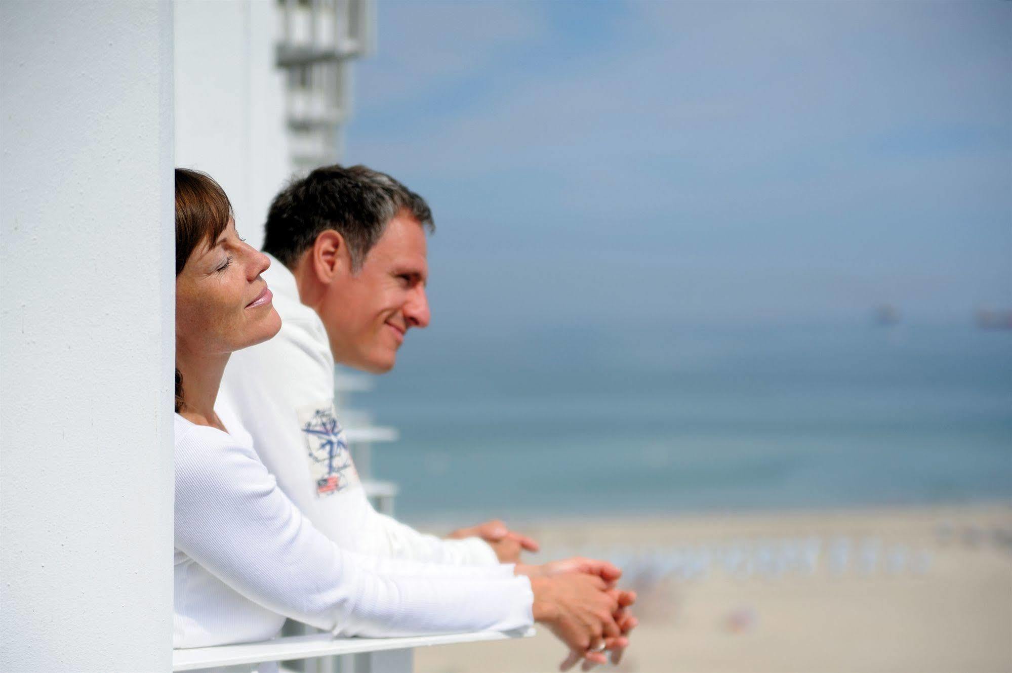 Hotel Neptun Rostock Exterior photo The photo depicts a man and a woman relaxing on a balcony. They appear to be enjoying a moment together, with the woman leaning back and closing her eyes, while the man looks forward with a smile. The background features a serene beach scene with oce