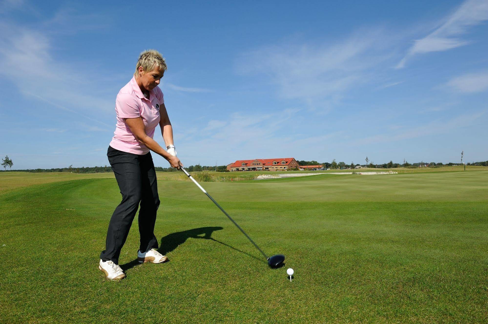 Hotel Neptun Rostock Exterior photo The image shows a person on a golf course preparing to take a shot. The individual is wearing a pink polo shirt and black pants, holding a golf club with both hands. In front of them, there is a golf ball teed up on the grass. The background features