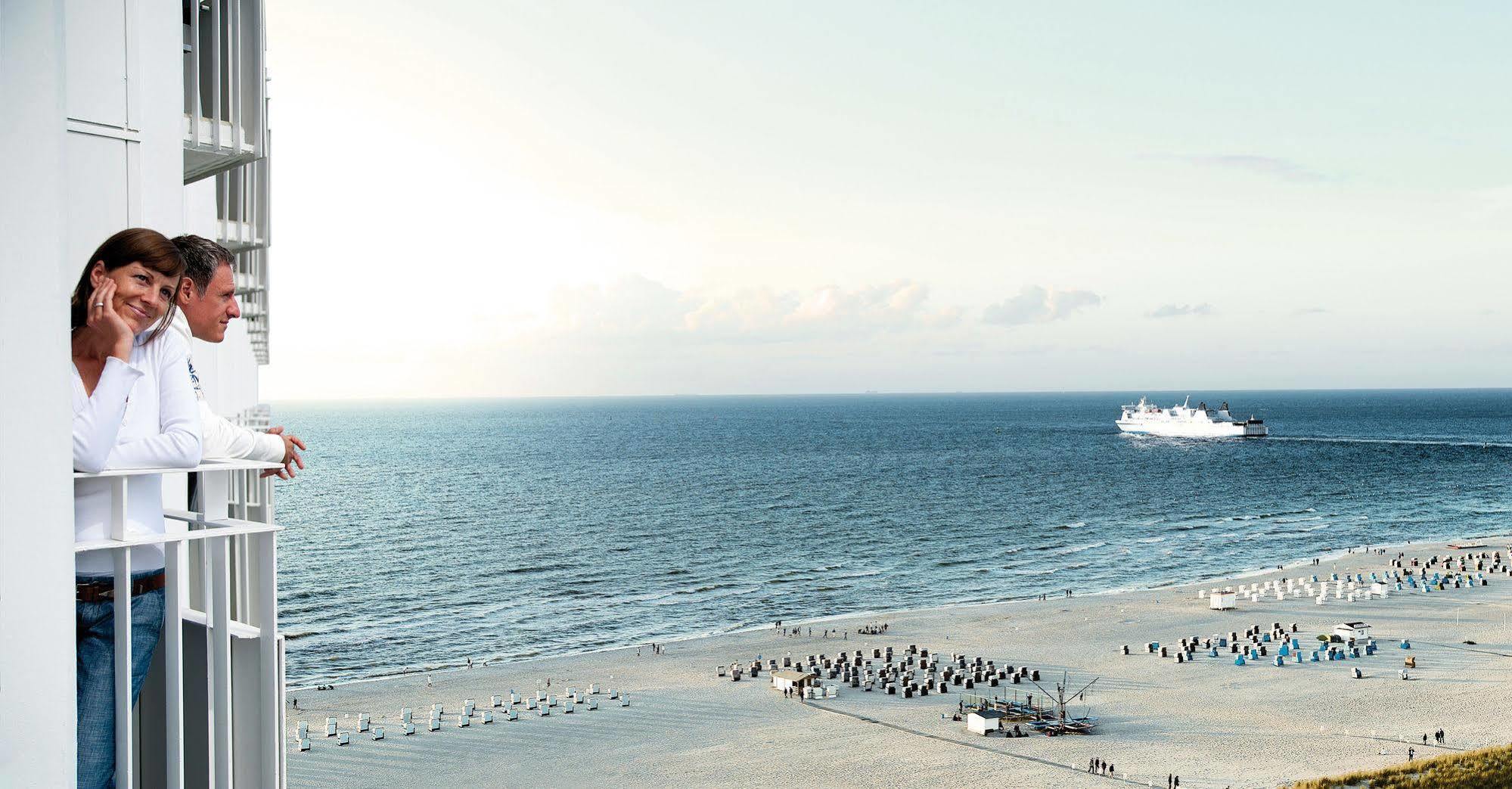 Hotel Neptun Rostock Exterior photo The image shows a coastal scene with a beach and ocean. In the foreground, there's a sandy beach with a group of people sitting under umbrellas. The ocean stretches out towards the horizon, and a ship is visible on the water. In the background, there