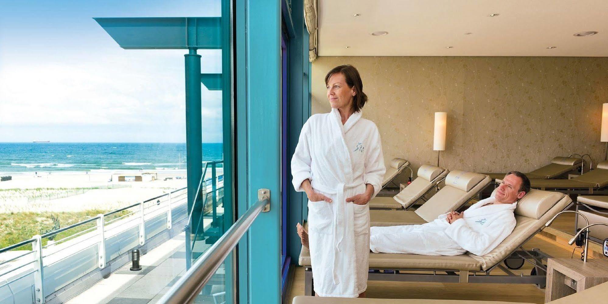 Hotel Neptun Rostock Exterior photo The photo shows a woman in a white bathrobe standing near a large window with a view of the ocean. She appears to be relaxed and contemplative, looking out at the water. In the background, there are lounge chairs, and another person is lying down, su