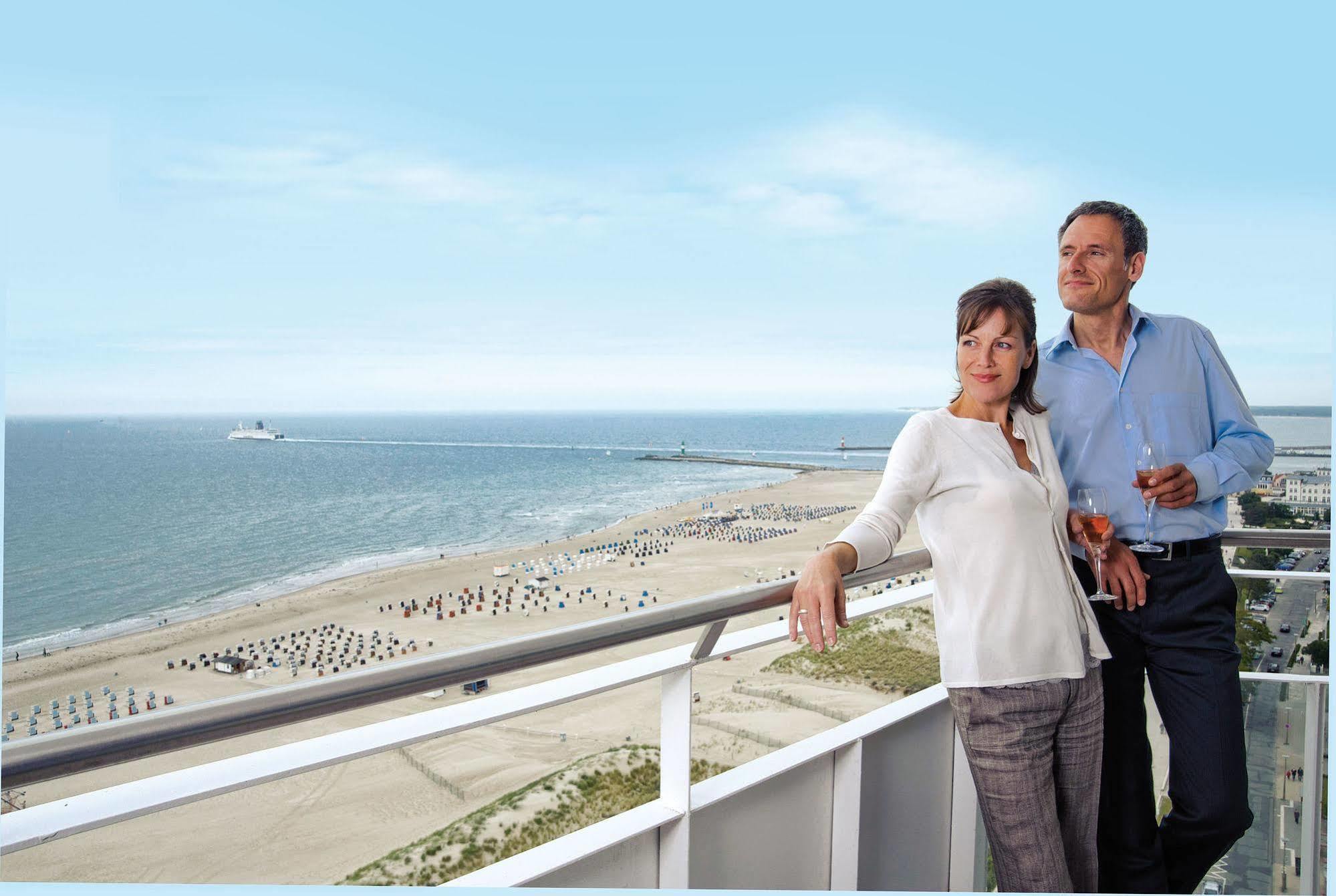 Hotel Neptun Rostock Exterior photo The photo shows a couple standing on a balcony overlooking a beach. They appear to be enjoying a scenic view of the ocean and coastline. The beach has some umbrellas or beach chairs arranged in rows, and the sky is clear with a bright blue tone. The 