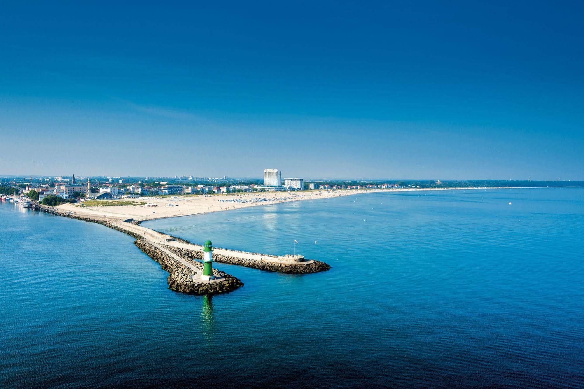 Hotel Neptun Rostock Exterior photo The photo showcases a serene coastal scene. In the foreground, there is a jetty extending into calm, blue waters, marked by a small green lighthouse at its end. The background features a sandy beach with a clear sky overhead, indicating a sunny day. 