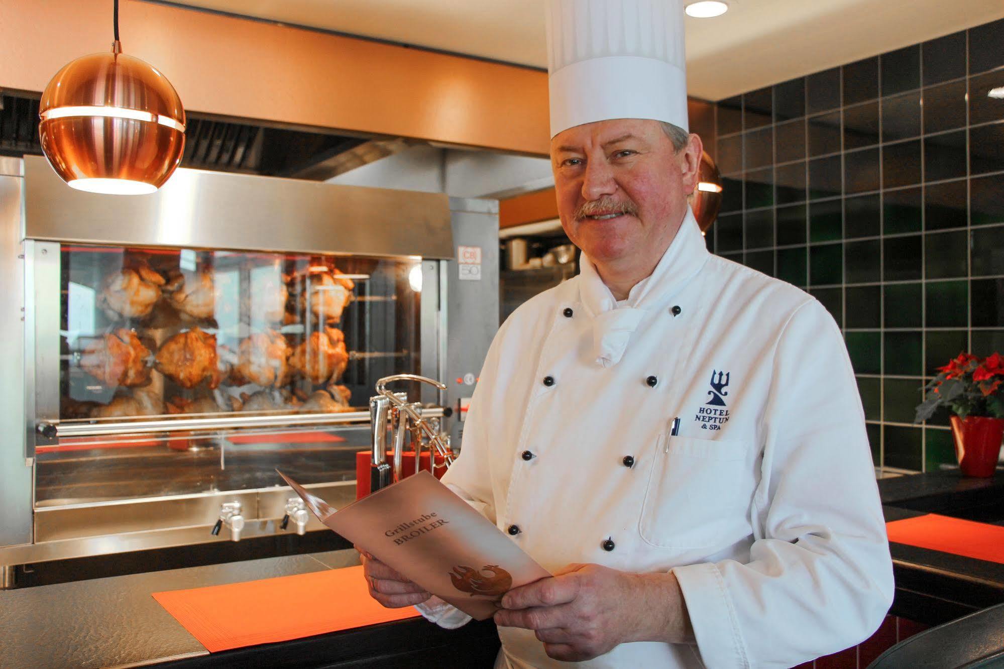 Hotel Neptun Rostock Exterior photo The photo shows a chef standing in a restaurant kitchen. He is wearing a white chef's uniform and a tall chef hat. The chef is holding a menu or a dish list and smiling. Behind him, there is a large oven or grill with roasted items visible, likely co