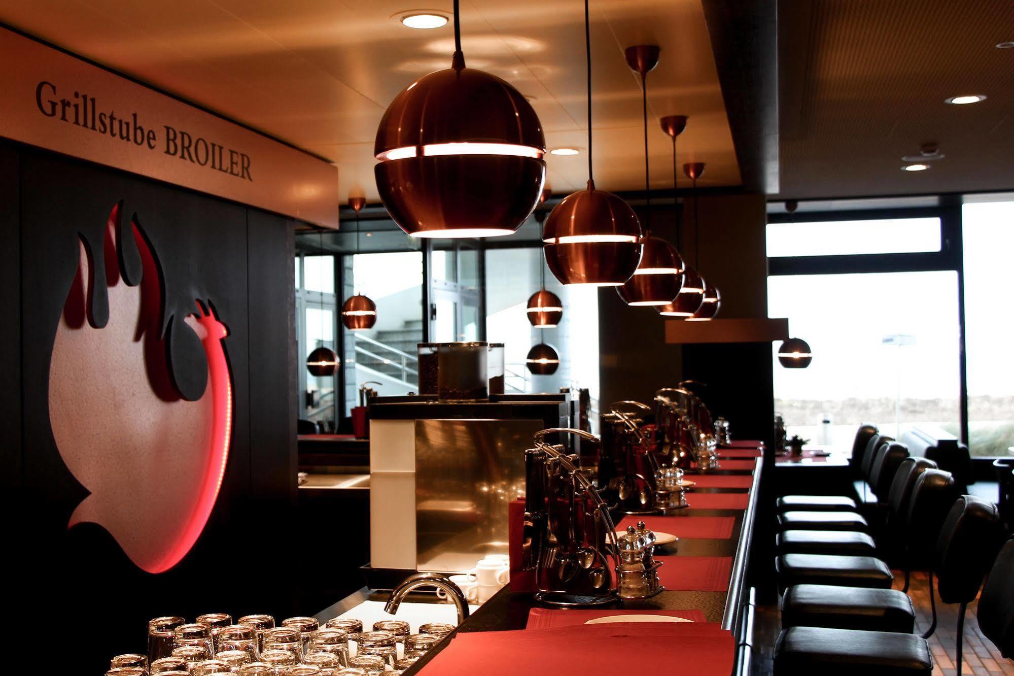 Hotel Neptun Rostock Exterior photo The photo depicts the interior of a modern dining establishment, likely a restaurant or grill. The design features a long bar or counter with a red surface, complemented by black seating. Pendant lights with a circular design hang from the ceiling, p