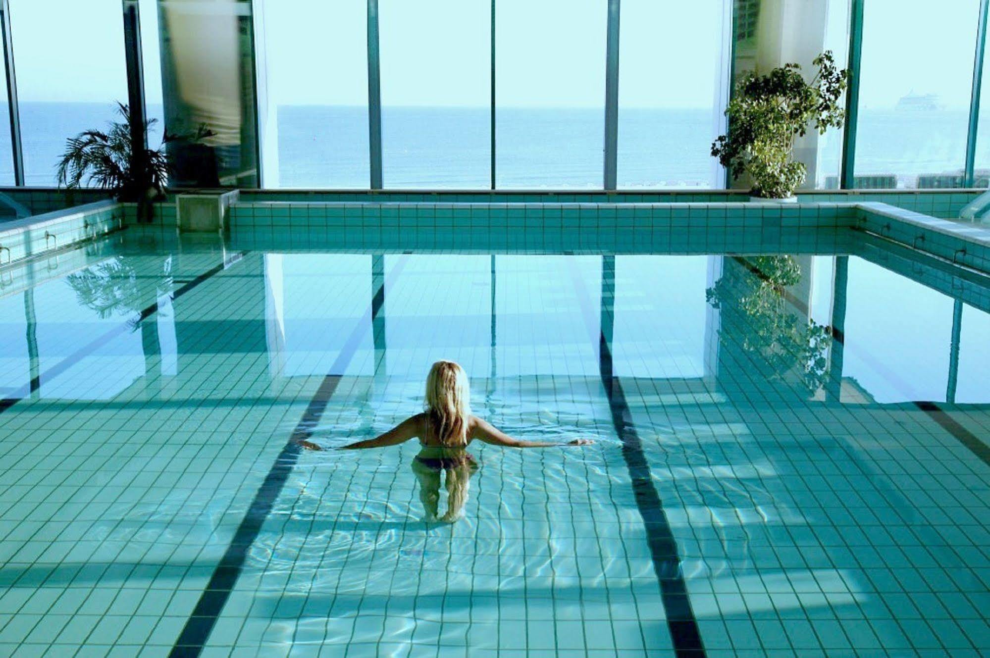 Hotel Neptun Rostock Exterior photo The image depicts an indoor swimming pool with large windows that overlook a scenic view of the ocean. A person is seen swimming or floating in the pool, facing away from the camera. The water is clear and reflects the light, creating a serene atmosp