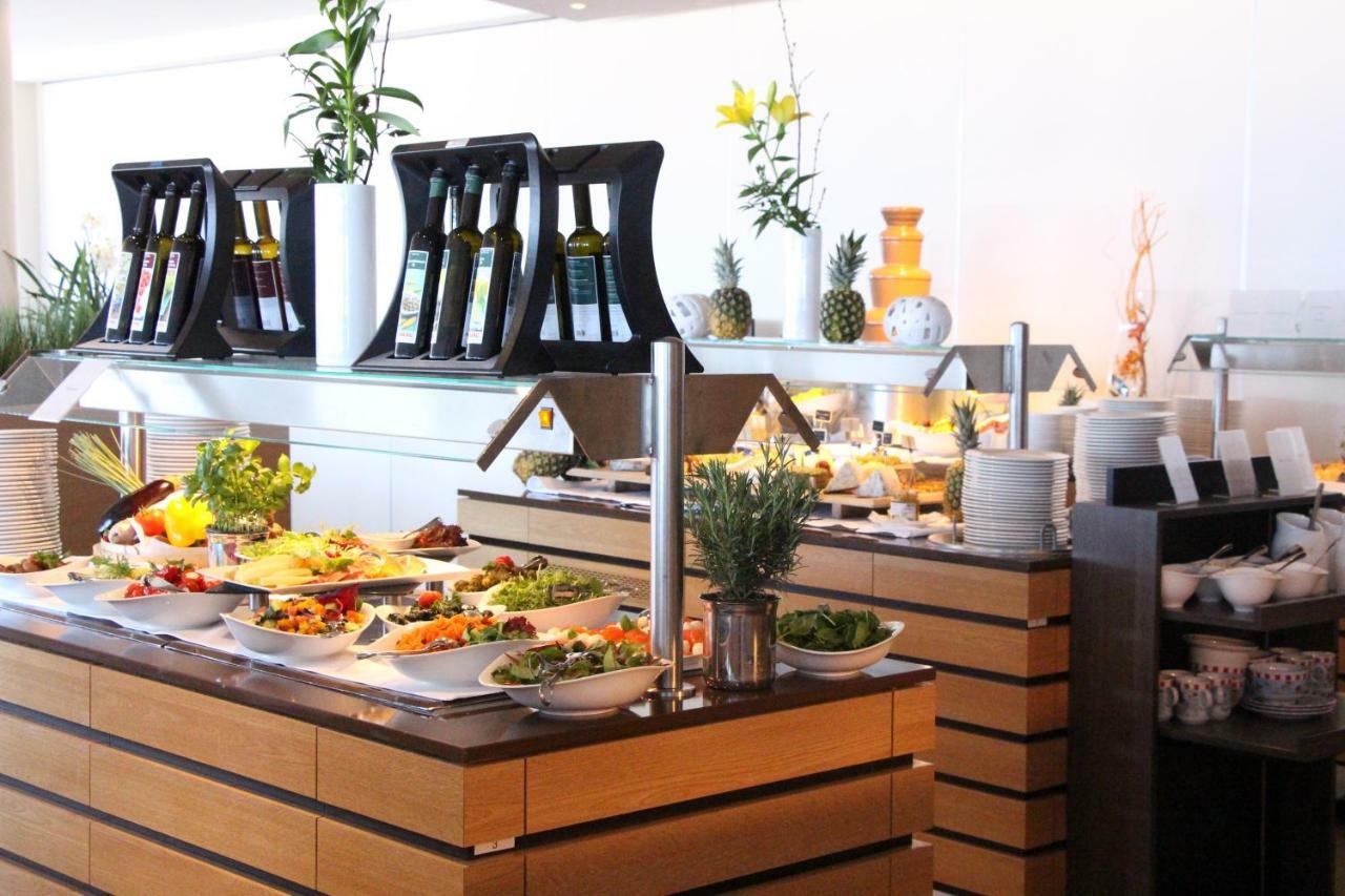 Hotel Neptun Rostock Exterior photo The photo shows a buffet-style food display with a variety of dishes arranged on a wooden counter. There are several plates filled with colorful salads and vegetables, indicating a fresh and healthy selection. Behind the food, there are bottles of oi