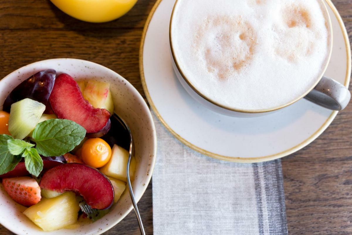 Hotel Neptun Rostock Exterior photo The photo shows a bowl of fresh fruit and a cup of cappuccino. The fruit bowl contains various pieces of fruit, including slices of red plums, melon, strawberries, and possibly a few other fruits, garnished with a mint leaf. Next to the fruit, there'