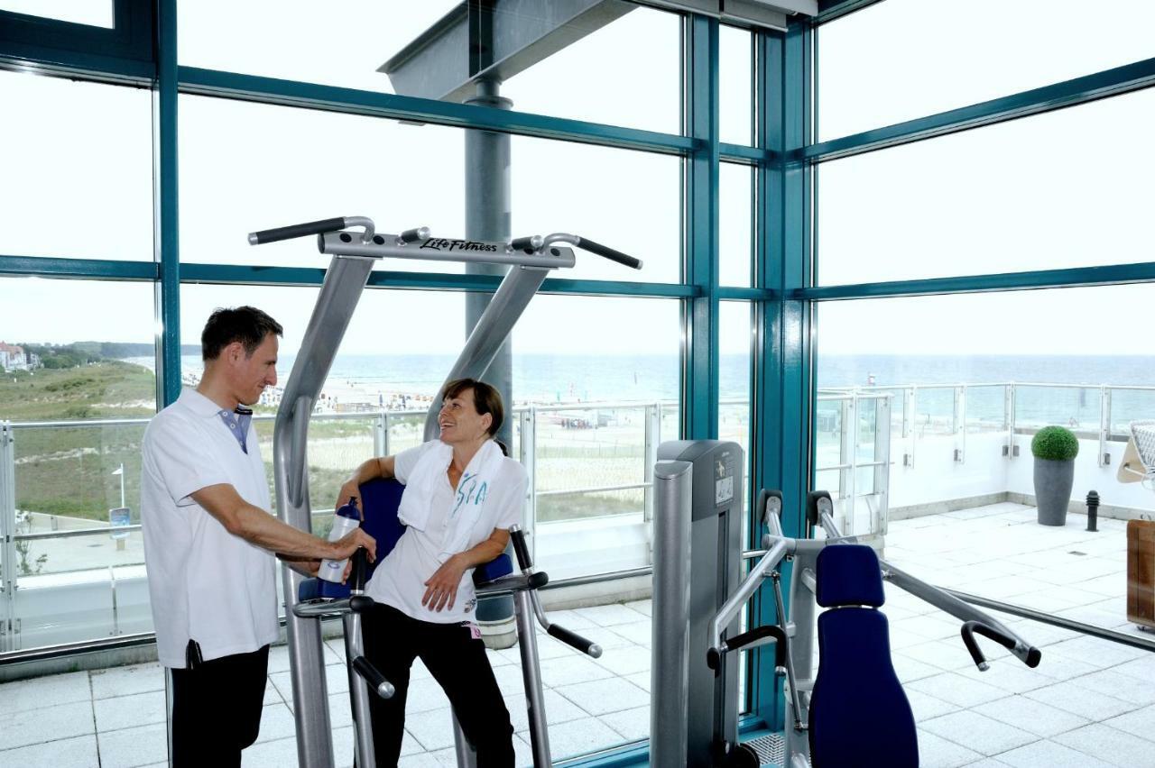 Hotel Neptun Rostock Exterior photo The photo shows two individuals in a bright, modern gym setting with large windows overlooking a view. One person is using a piece of gym equipment, possibly a weight machine, while the other person stands nearby, possibly providing assistance or gui