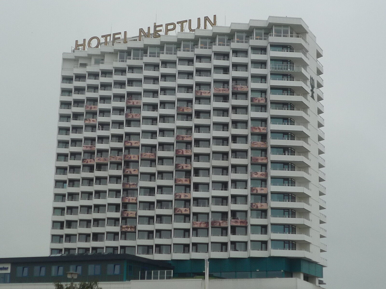 Hotel Neptun Rostock Exterior photo The photo shows a tall, modern hotel building named "Hotel Neptun." It has a white facade with multiple rectangular windows and balconies. The top of the building features the hotel name prominently displayed. The sky appears overcast, indicating clo