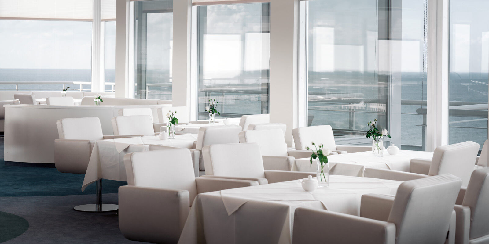 Hotel Neptun Rostock Exterior photo The photo shows a modern dining area with multiple tables set up. The tables are covered with white tablecloths and have small vases with flowers as centerpieces. The seating consists of white chairs that are simple and contemporary in design. Large 